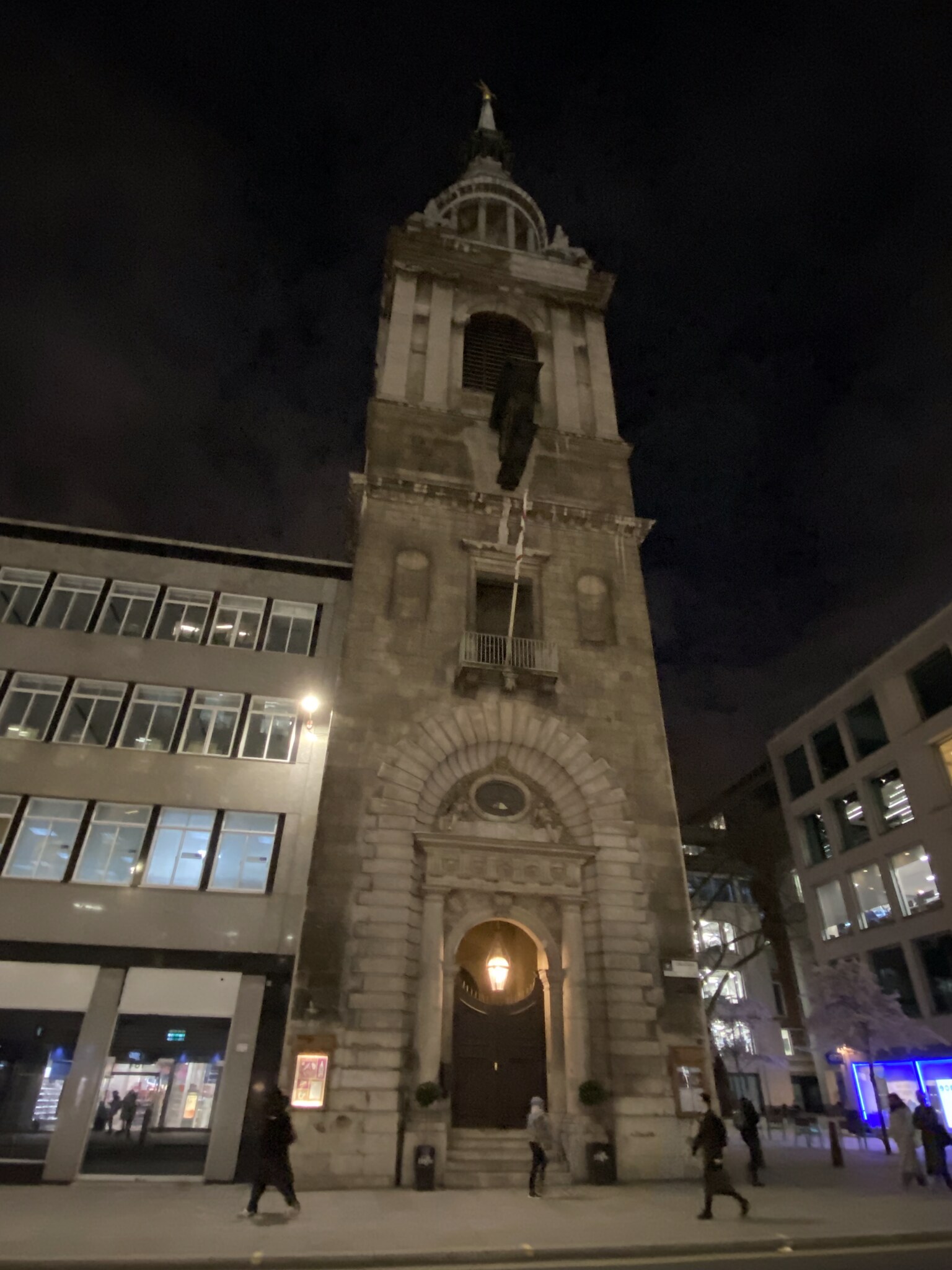 Een Wren-kerktoren verlicht in de avond in de City of London, omgeven door donkere Victoriaanse straatjes