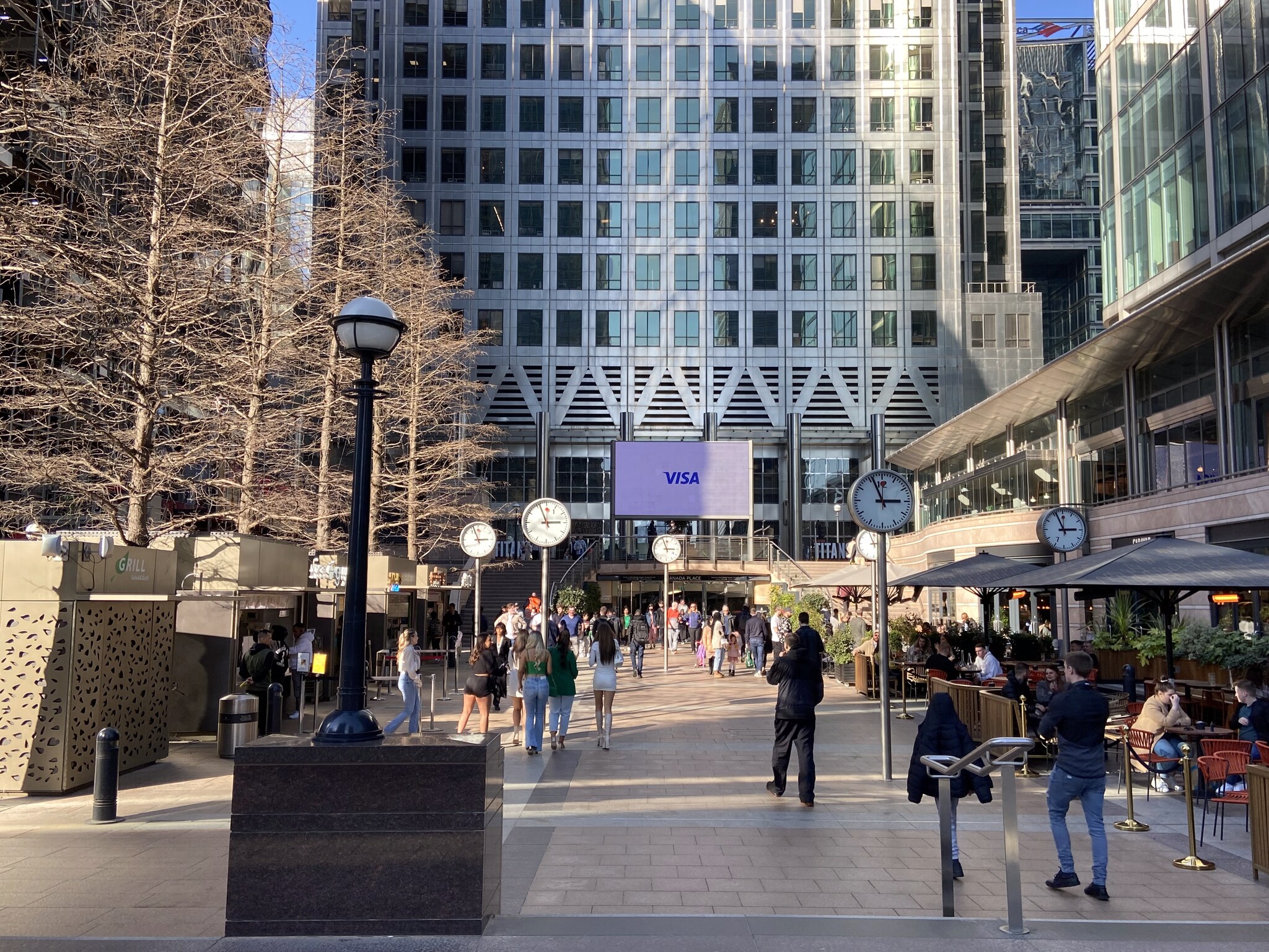 De bruisende plaza van Canary Wharf met winkels, terrassen en de glazen kantoortorens op de achtergrond op een zonnige dag
